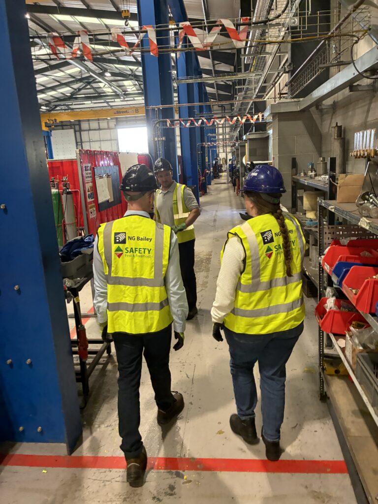 Three workers wearing yellow safety vests and hard hats walk through an industrial facility with tools and equipment along the sides. One worker faces forward, while the other two have their backs to the camera.