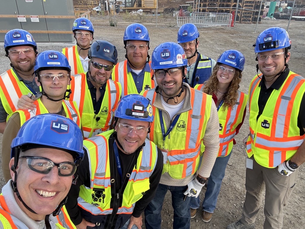 A group of twelve construction workers wearing blue hard hats, safety glasses, and high-visibility vests stand together outdoors at a construction site, smiling for a group photo.