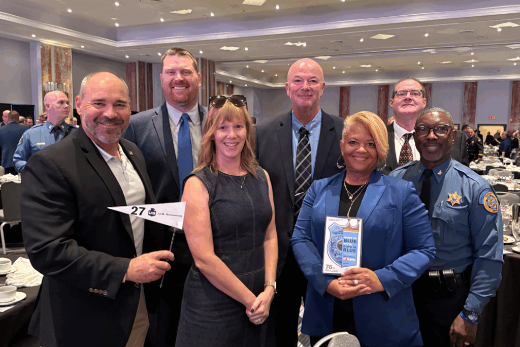 Seven adults, four men in suits, one man in a police uniform, and two women in dresses, stand together posing and smiling at a formal event with round tables and other attendees visible in the background.