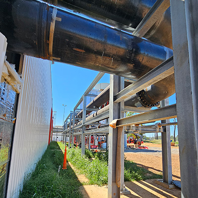 Large black industrial pipes are mounted on a metal support structure next to a corrugated white building, with grass and dirt visible below and construction equipment in the background under a clear blue sky.