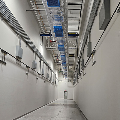 A long, empty industrial hallway with white walls, exposed piping, and ducts covered in silver insulation with blue filters on the ceiling; a closed gray door is at the far end of the corridor.