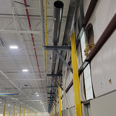 Large metal pipes are mounted on yellow supports along the ceiling and wall of an industrial warehouse. The wall has a large hole with exposed insulation, and glass blocks are visible nearby. The area is brightly lit.