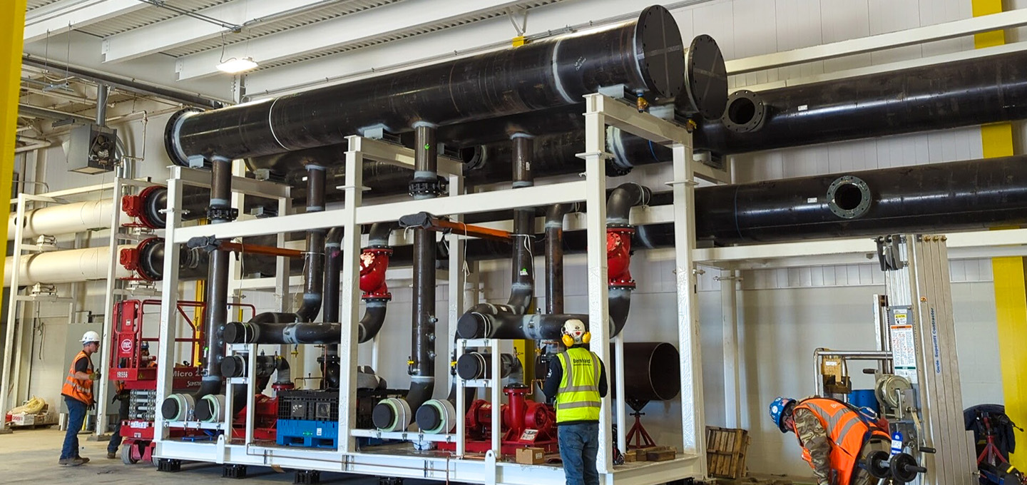 Several workers in safety gear stand near a large industrial piping system with black pipes, red valves, and white structural supports inside a spacious, well-lit facility.