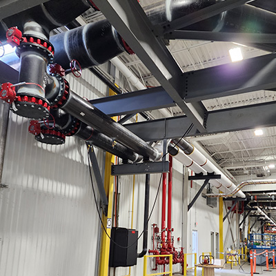 Industrial piping with red valve handles runs along a wall and ceiling inside a large facility. Metal beams support the pipes, and fire suppression equipment is visible in the background. The area is well-lit.