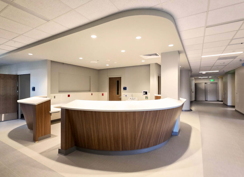 A modern, empty hospital or clinic reception desk with a wood finish and white countertop is situated in a clean, well-lit hallway with doors and overhead lighting visible in the background.