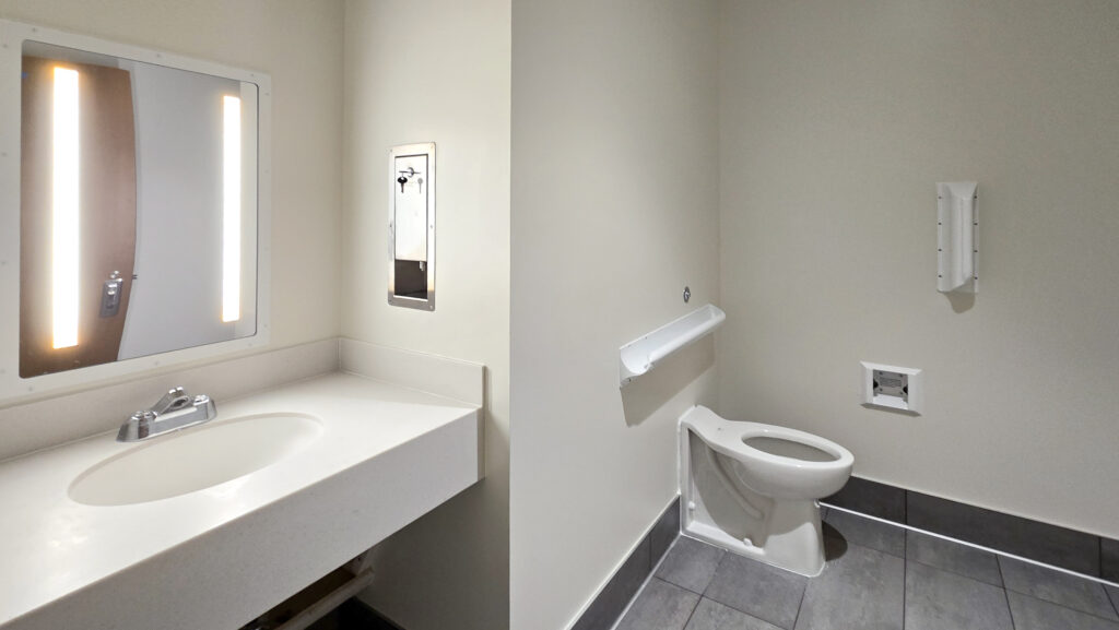 A public restroom with a sink, lit mirror, and countertop on the left, and a toilet with handrails and tiled floor on the right. The walls are light-colored and the room appears clean and accessible.