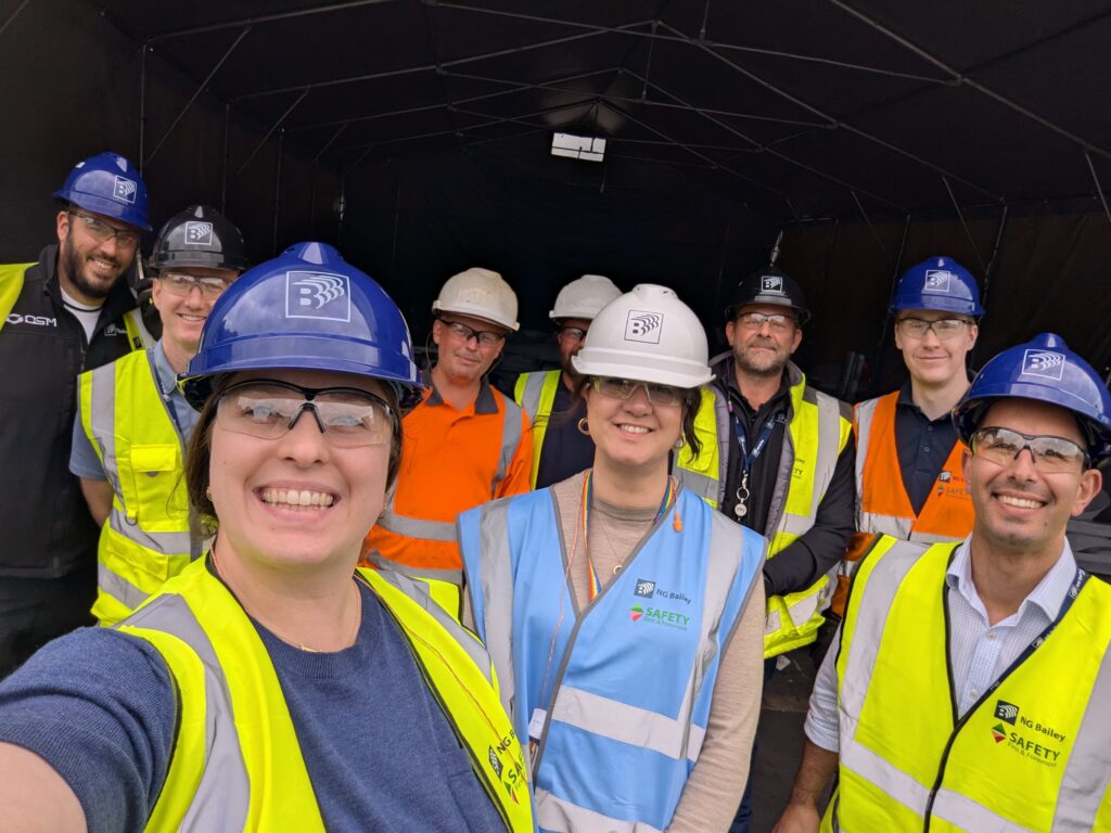 A group of ten people wearing safety helmets, reflective vests, and safety glasses poses for a photo inside a large covered work area. Some are smiling, and two people in front are taking the selfie.