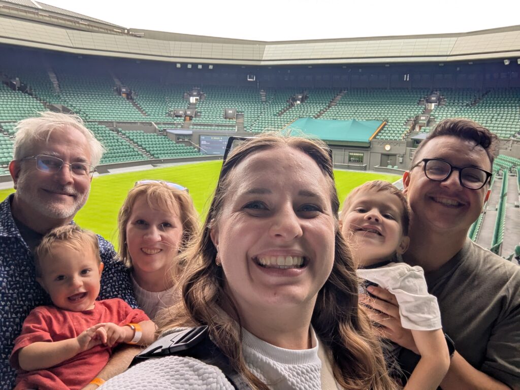 A group of six people, including two young children, smiles for a selfie in a stadium with green seating and a grass court, likely at Wimbledon. The stadium appears mostly empty in the background.