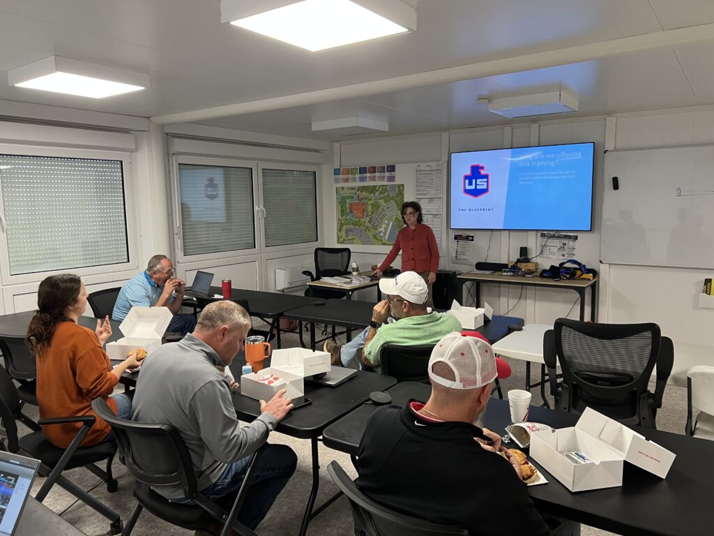 A group of people sit at tables eating boxed meals while a woman stands at the front of the room giving a presentation. A screen displays a slide with a logo and text. Office supplies and notebooks are on the tables.