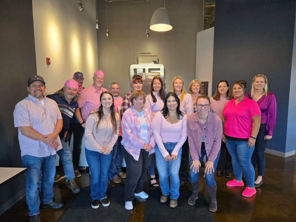 A group of sixteen adults stands indoors, posing for a photo. Most are wearing pink or light-colored shirts. They are smiling and standing in front of a gray wall with a logo. The lighting is bright.