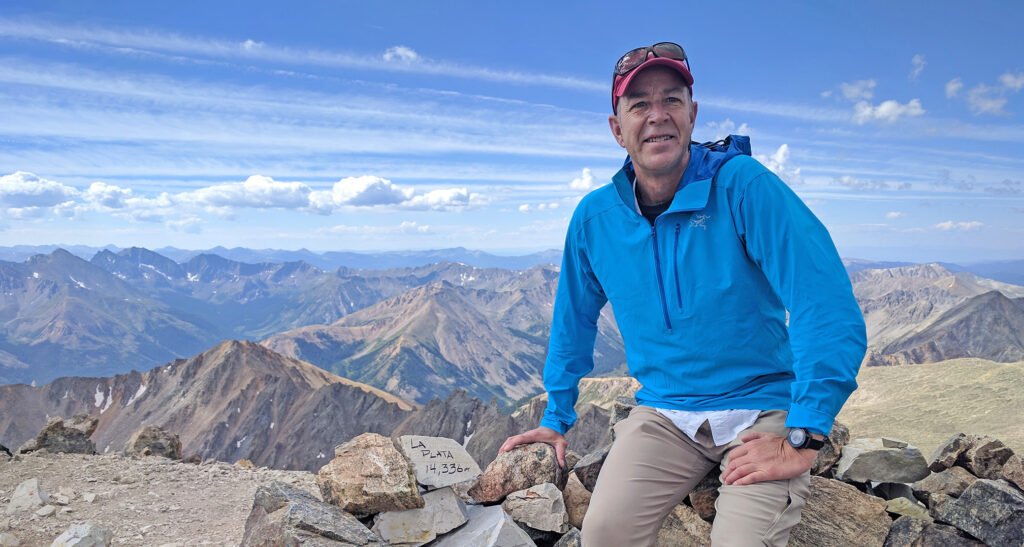 A man in a blue jacket and red cap poses on a rocky mountain summit with a sign reading “La Plata 14,336.” Mountain peaks and a partly cloudy sky are visible in the background.