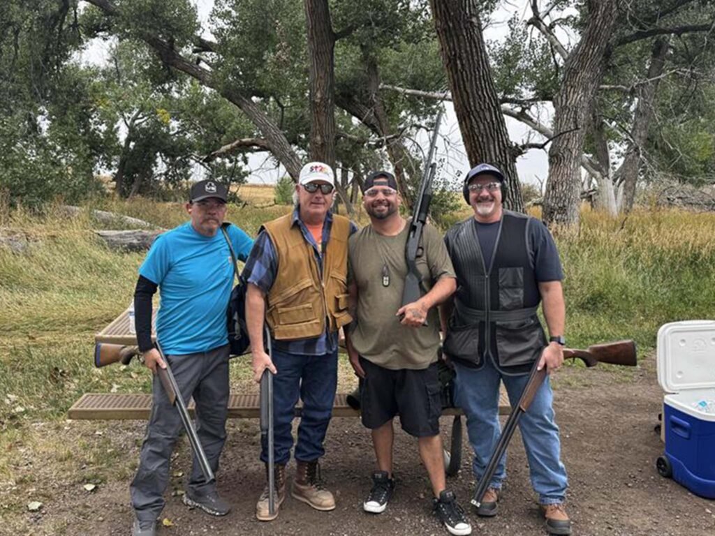 Four men stand outdoors on dirt ground in front of trees, each holding a shotgun and wearing casual clothes with protective vests. A metal picnic table and a blue cooler are visible in the background.