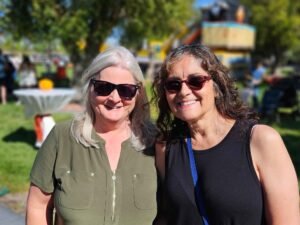 Two women stand close together outdoors on a sunny day, both wearing sunglasses and smiling. There are trees, grass, and people in the background, suggesting a park or outdoor event.