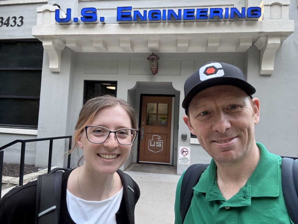 Two people with backpacks stand outside a building entrance with a sign above that reads U.S. Engineering. The man wears a black cap and green shirt; the woman wears glasses and a backpack.