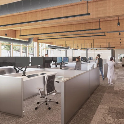 A modern open-plan office with cubicles, multiple computer monitors, and ergonomic chairs. Several people are working or walking around. The space features wooden ceilings, exposed ductwork, and large windows.