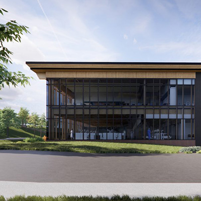 A modern two-story building with large glass windows, surrounded by greenery and a paved road in front. The sky is mostly clear with a few wispy clouds.