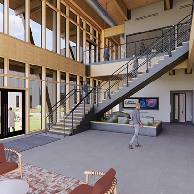 A modern building lobby with large windows, a central staircase with metal railings, indoor plants, chairs, a rug, and people walking or standing on both floors. Walls and ceilings feature wood accents.