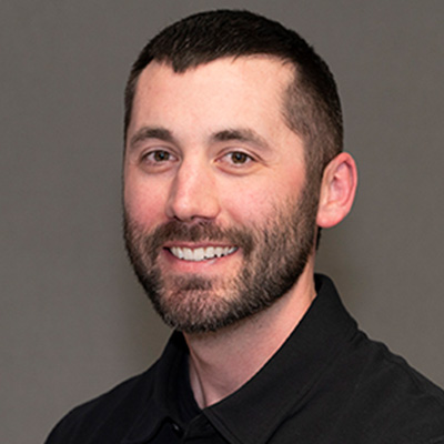 A man with short dark hair and a beard, wearing a black collared shirt, smiles at the camera against a plain gray background.