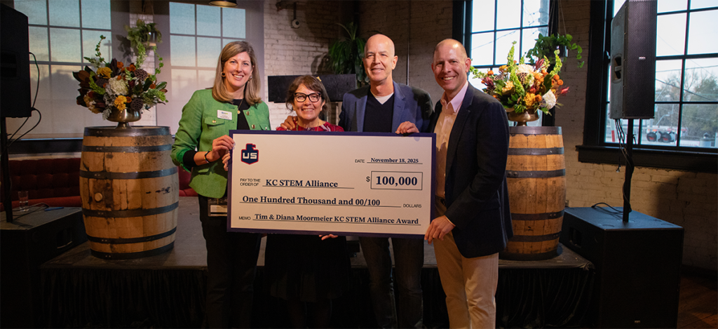 Four people stand indoors, smiling and holding a large ceremonial check for $100,000 made out to KC STEM Alliance. Two wooden barrels with flower arrangements are visible in the background.