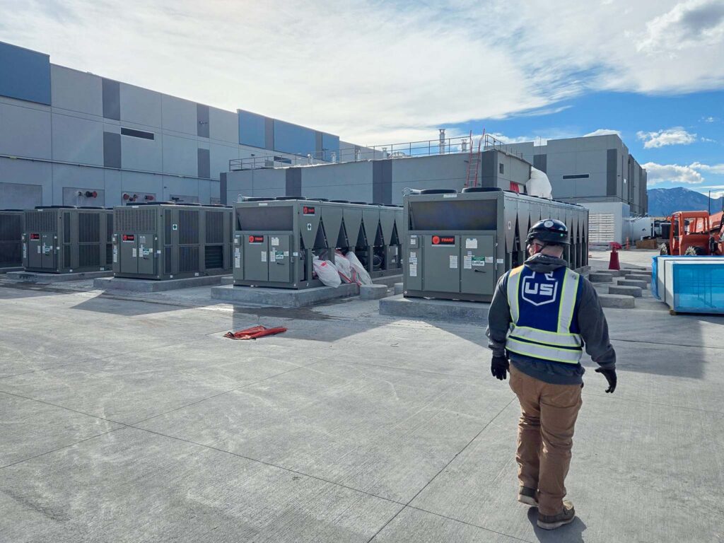 A worker wearing a helmet and safety vest walks on a concrete lot near several large HVAC units outside a grey industrial building under a partly cloudy sky.