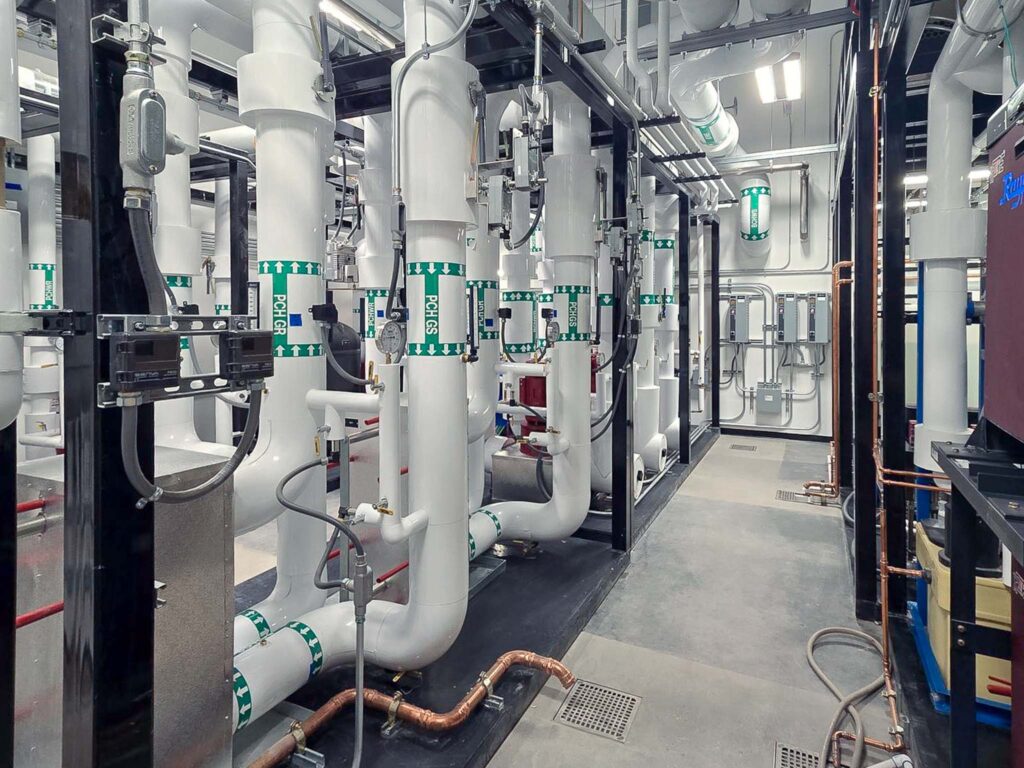 Room with large white industrial pipes labeled “CHWS,” metal framework, exposed valves, copper piping, and ceiling conduits. Overhead fluorescent lights illuminate the clean, modern utility area.