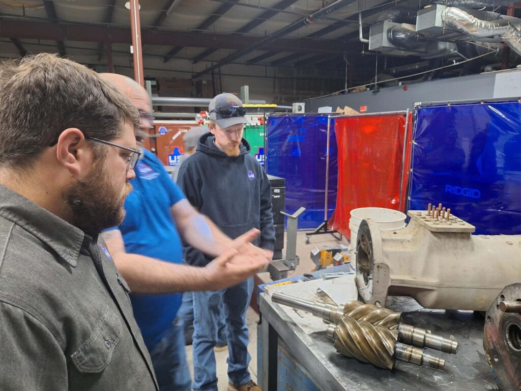Three men stand around a workbench in an industrial workshop, examining a large mechanical component and gears. Colorful welding screens and various equipment are visible in the background.
