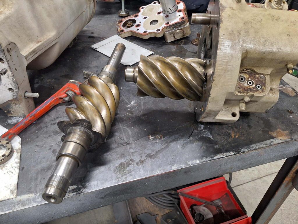 Two metal helical rotors, one removed and one partially installed in a mechanical housing, are on a workshop table with various tools, parts, and papers scattered around.