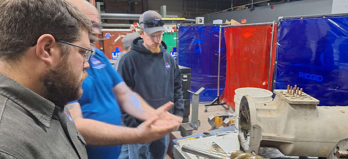 Three men stand in a workshop, two focused on a metal machine part on a workbench while one gestures with his hands. The background includes red and blue welding screens and various equipment.