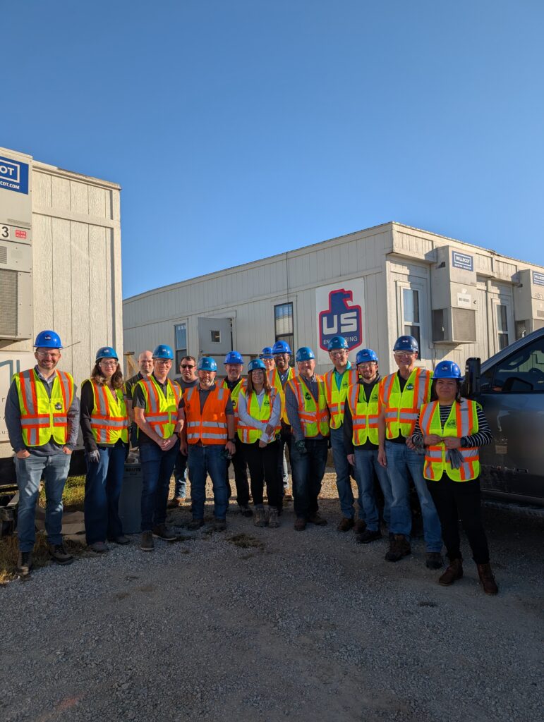 A group of fifteen people wearing blue hard hats and yellow safety vests stands on gravel outside portable office buildings with “US” and other signage visible in the background. The sky is clear and sunny.