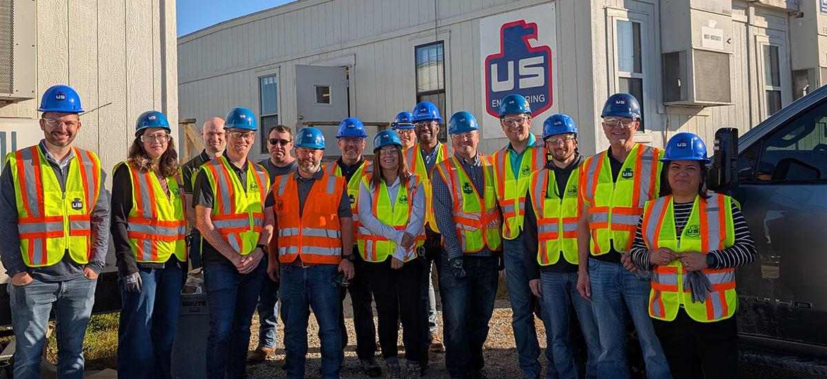 A group of people wearing blue hard hats and orange or yellow safety vests stand together outside in front of a building labeled “US Engineering” on a sunny day.