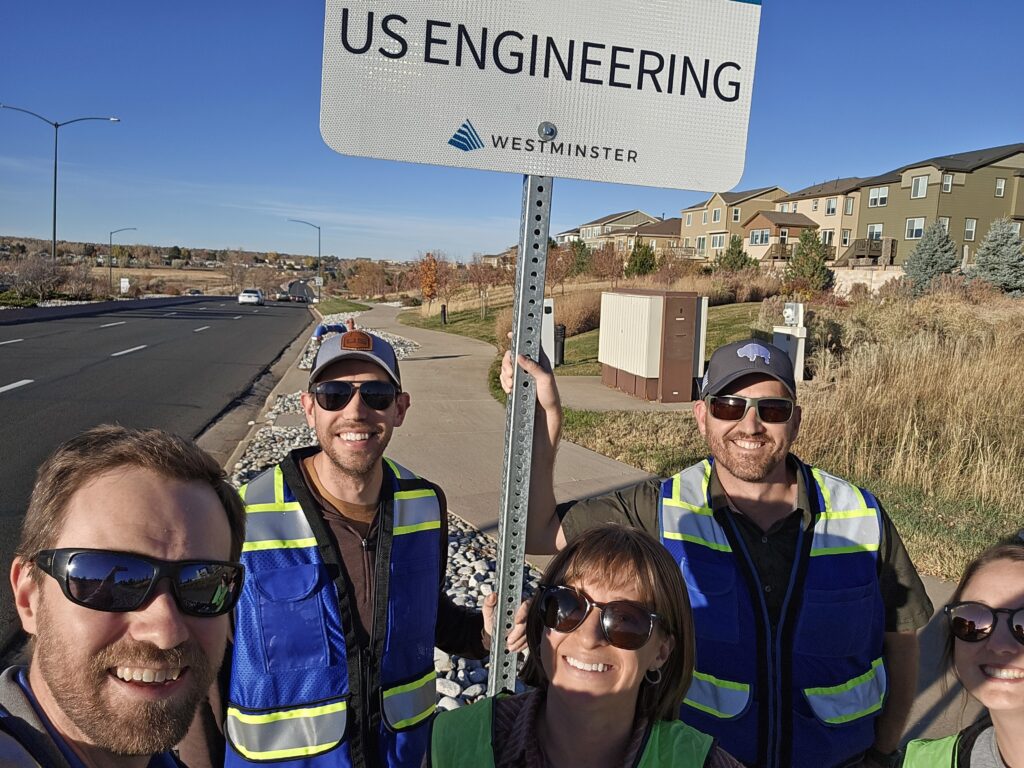 Five people wearing safety vests and sunglasses stand outdoors on a sidewalk next to a road, smiling at the camera. They are holding a sign that reads US ENGINEERING WESTMINSTER. Houses are visible in the background.