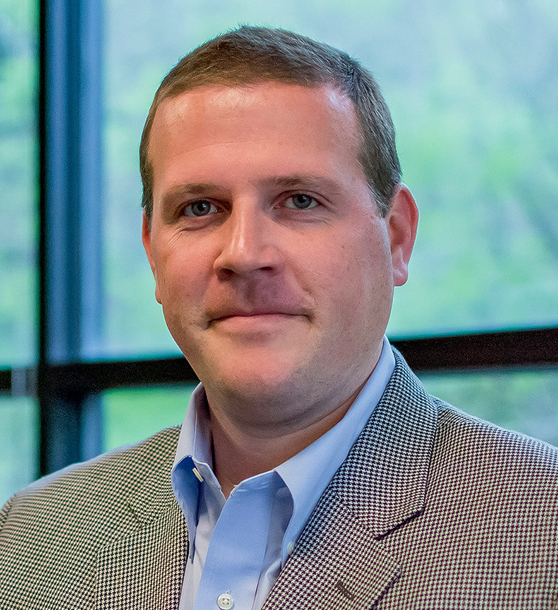 A man with short light brown hair wearing a light blue dress shirt and a gray checked blazer stands indoors in front of large windows with a blurred green outdoor background.