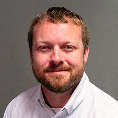A man with short light brown hair, a beard, and a mustache is smiling softly. He is wearing a white collared shirt and is posed against a plain gray background.