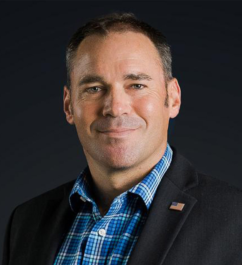 A man wearing a dark blazer with an American flag pin and a blue checked shirt stands against a dark background, looking at the camera with a slight smile.