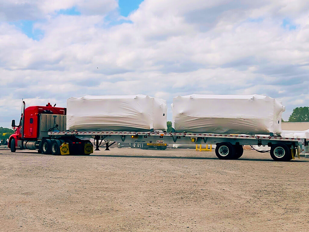 A red semi-truck with a flatbed trailer is parked on a gravel lot, transporting two large, white-wrapped rectangular objects secured to the trailer. The sky is cloudy with some blue patches visible.