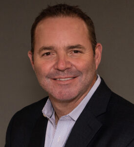 A middle-aged man with short brown hair, light stubble, and a slight smile is wearing a dark suit jacket and a light striped collared shirt. He is posing in front of a plain, neutral background.