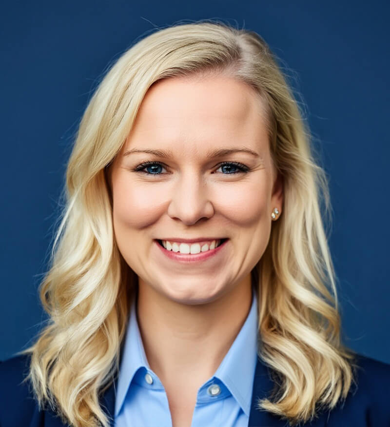 A woman with shoulder-length blonde hair smiles at the camera. She is wearing a light blue collared shirt and a dark blazer. The background is a solid dark blue.