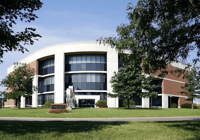A modern, three-story office or academic building with large glass windows and red brick accents, surrounded by green lawn, trees, and a small sign and sculpture in front.