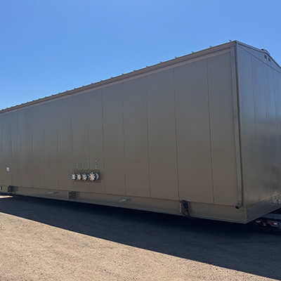 A large, rectangular, brown metal structure sits on a gravel surface outdoors under a clear blue sky. Four utility connections are visible on one side of the structure.