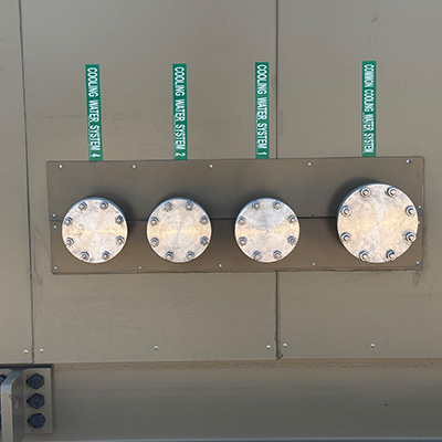Four metal circular access covers, each secured with multiple bolts, are mounted on a tan industrial panel. Above each cover, a vertical green label reads COOLING WATER SYSTEM 1 or COOLING WATER SYSTEM 2.