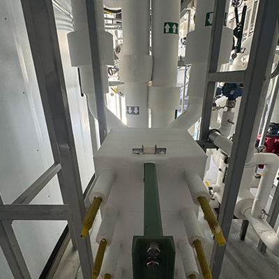 White industrial pipes and fittings are arranged vertically and horizontally inside a facility. Green signs with arrows, as well as various valves and connectors, are visible among the machinery. Lighting is moderate.