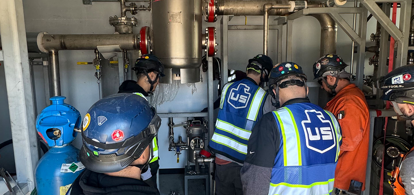 Several workers wearing safety gear, helmets, and vests are gathered around industrial equipment and pipes inside a facility. Some are observing closely while one appears to be operating or inspecting the machinery.
