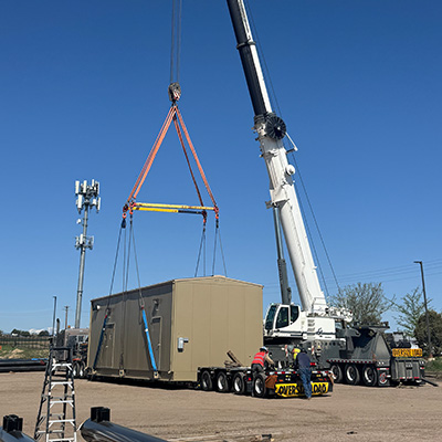A large crane lifts a beige modular building onto a flatbed trailer. Several workers in safety gear stand nearby. The scene takes place outdoors under a clear blue sky.