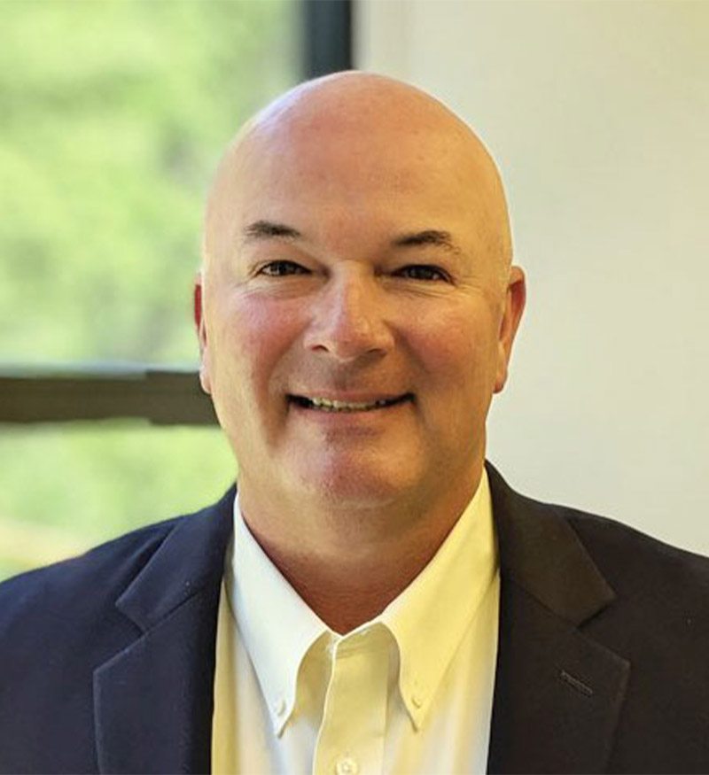 A middle-aged man with a bald head, wearing a white dress shirt and a dark blazer, is smiling at the camera. A window with a blurred green outdoor scene is in the background.