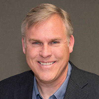 A smiling middle-aged man with short, light gray hair wearing a dark checked blazer over a blue collared shirt, standing in front of a neutral gray background.