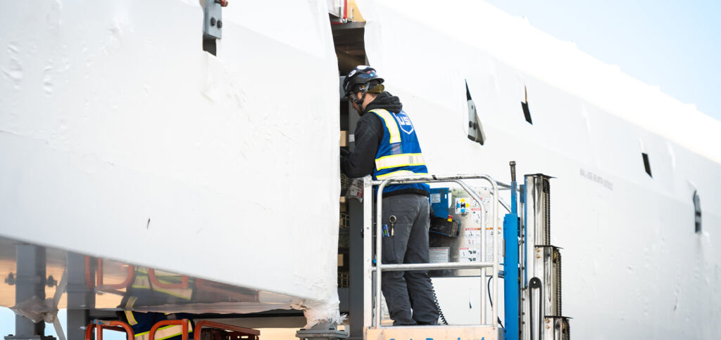 A worker in safety gear and a blue vest operates a lift while inspecting or working on a large, white industrial structure wrapped in protective material.