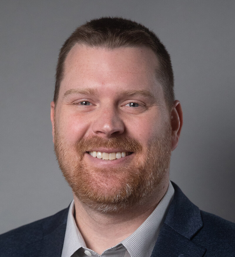 A man with short brown hair and a trimmed reddish beard is smiling. He is wearing a gray collared shirt and a dark jacket against a plain gray background.