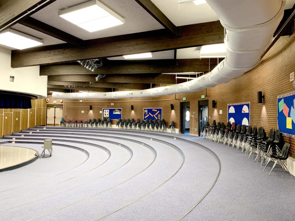 A large, empty auditorium with tiered, carpeted seating, stacked black chairs against a brick wall, blue bulletin boards with artwork, and a stage on the left side. Ceiling lights and exposed pipes are visible.