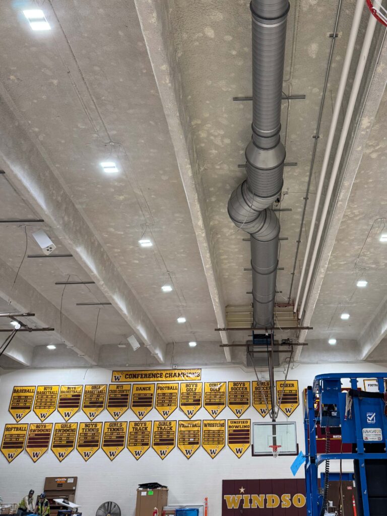 A gymnasium ceiling with exposed pipes and ducts, walls lined with yellow championship banners, a scoreboard, basketball hoops, and part of a blue lift visible in the foreground. The word Windsor is on the wall.