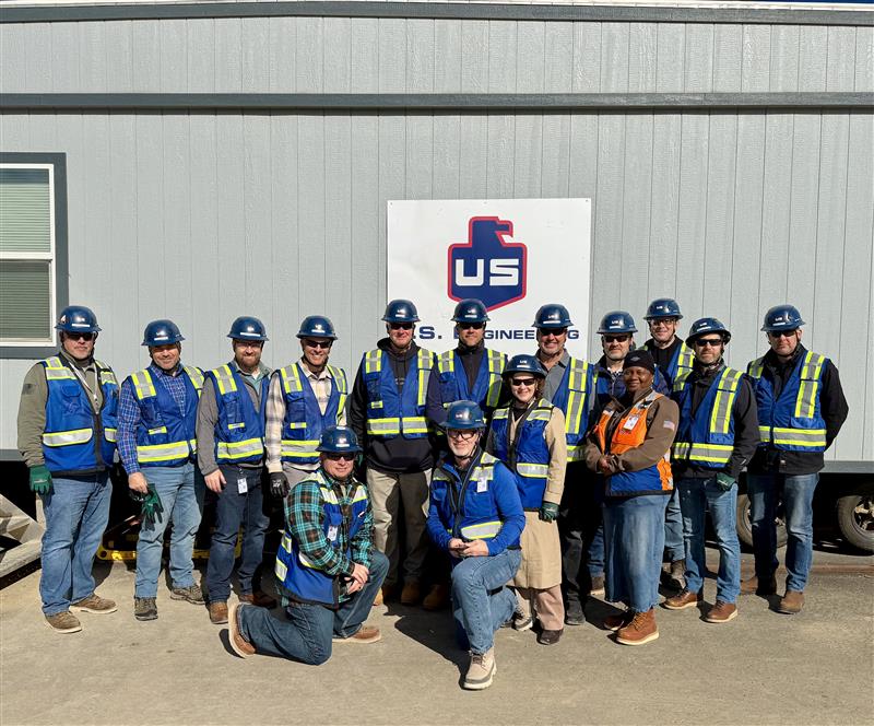 A group of fifteen construction workers wearing blue hard hats, safety vests, and work attire stand in front of a gray portable building with a U.S. Engineering sign.
